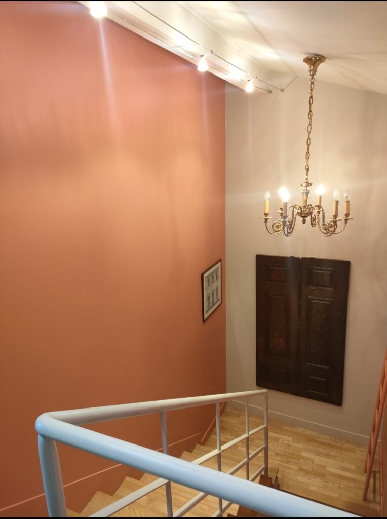 Escalier intérieur avec murs pêche et beige. Un chandelier doré, des spots et une rampe blanche. Cadre et portes en bois.