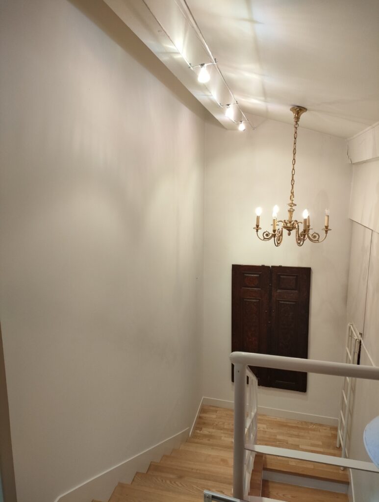 Escalier en bois clair menant vers un palier, éclairé par spots de plafond et un lustre doré ancien devant des portes sombres.