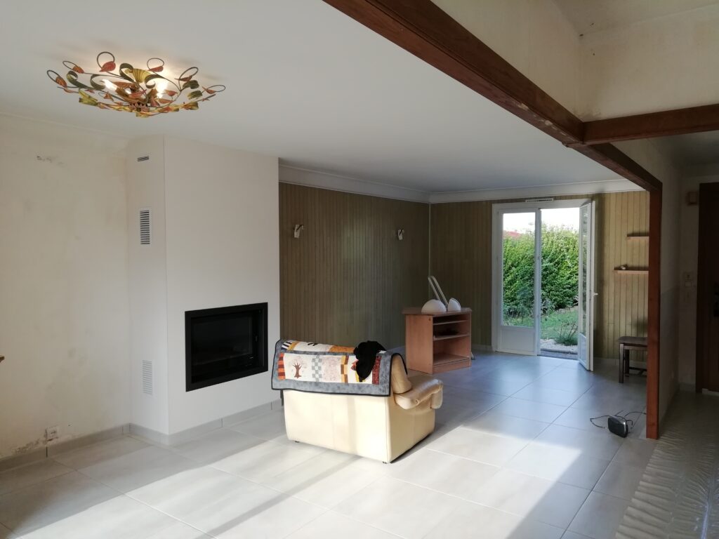 Salon rénové clair avec cheminée moderne noire, plafonnier floral, boiseries murales, carrelage blanc et porte vers jardin ensoleillé.