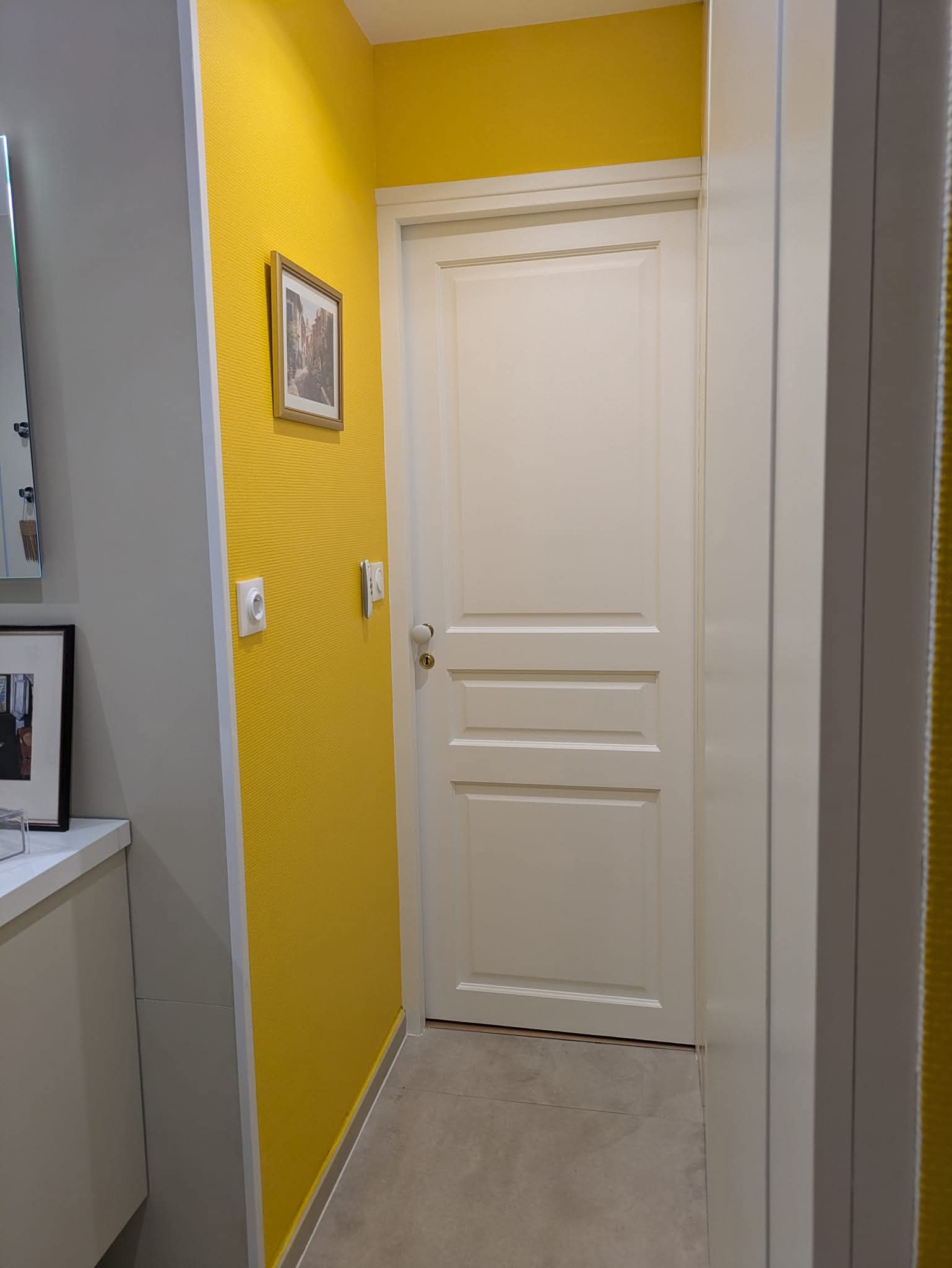 Couloir moderne avec mur jaune vif texturé, porte blanche à panneaux et petit cadre décoratif.
