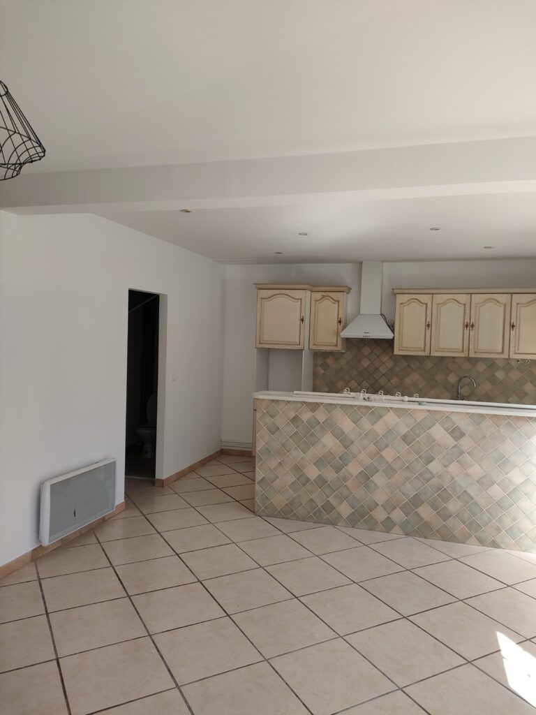 Cuisine ouverte avec îlot central carrelé, meubles en bois clair et carrelage au sol dans un appartement vide.