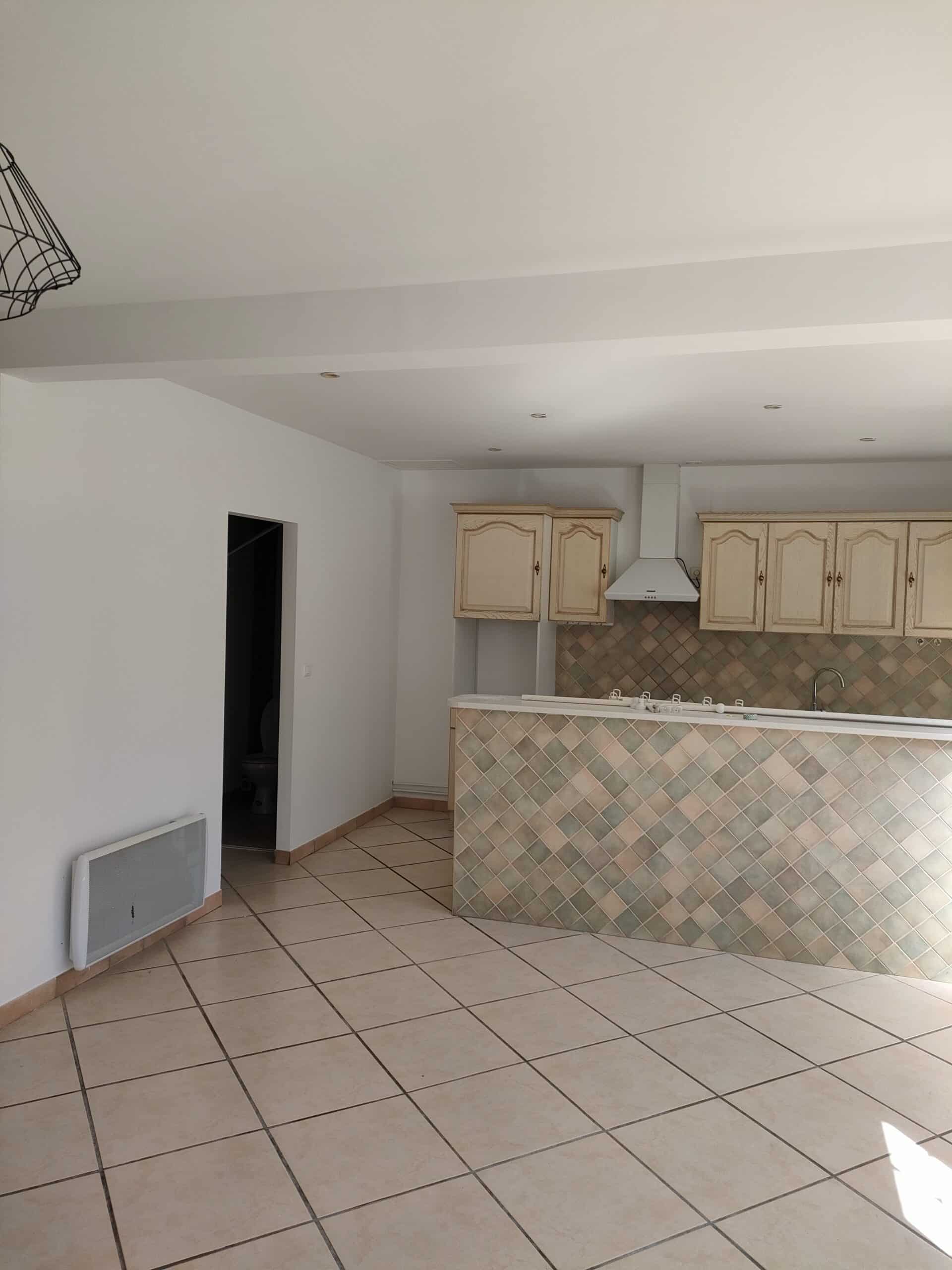 Cuisine ouverte avec îlot central carrelé, meubles en bois clair et carrelage au sol dans un appartement vide.