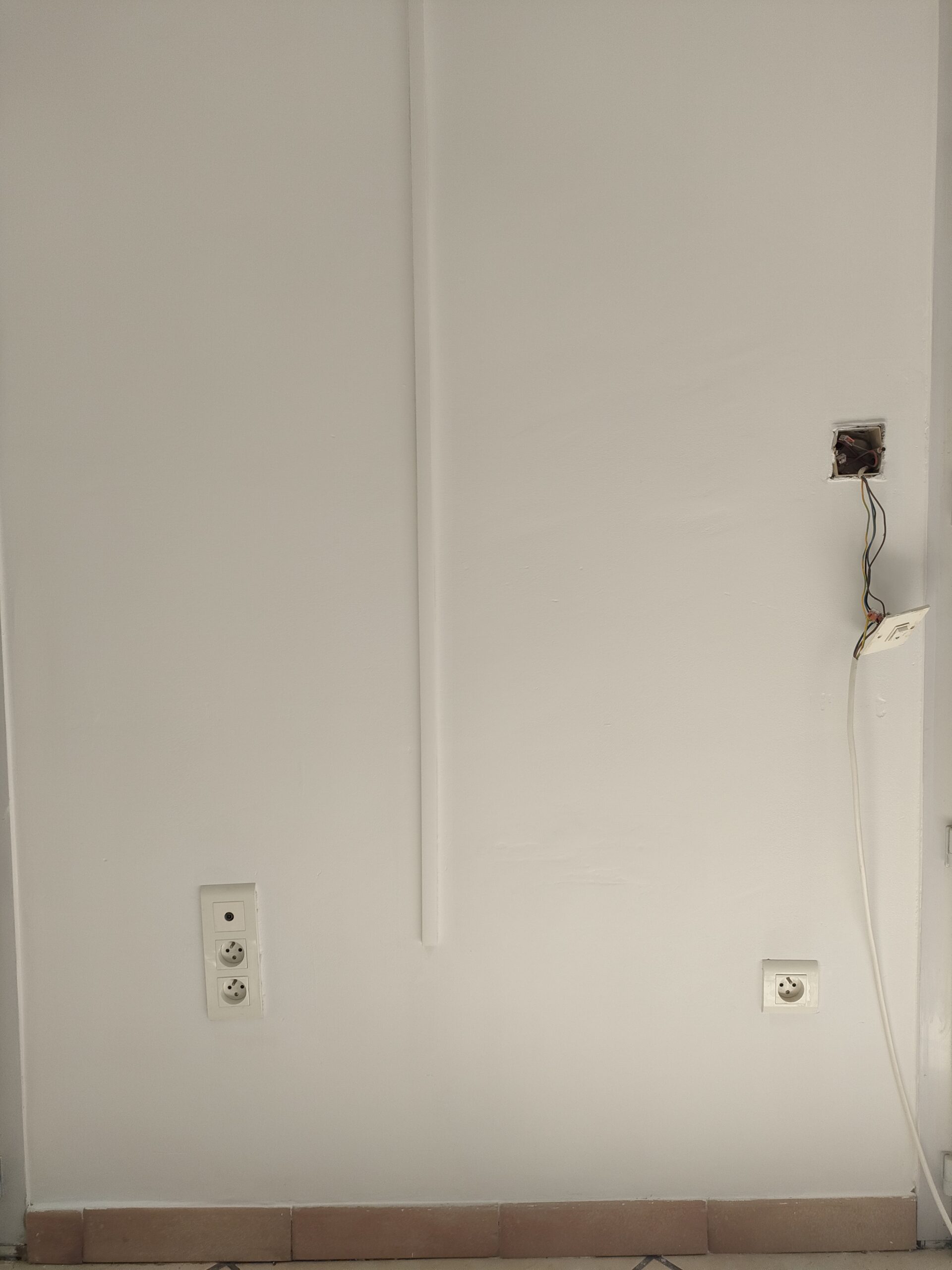 Mur blanc avec prises électriques, goulotte verticale et fils électriques apparents sortant d'un boîtier ouvert.