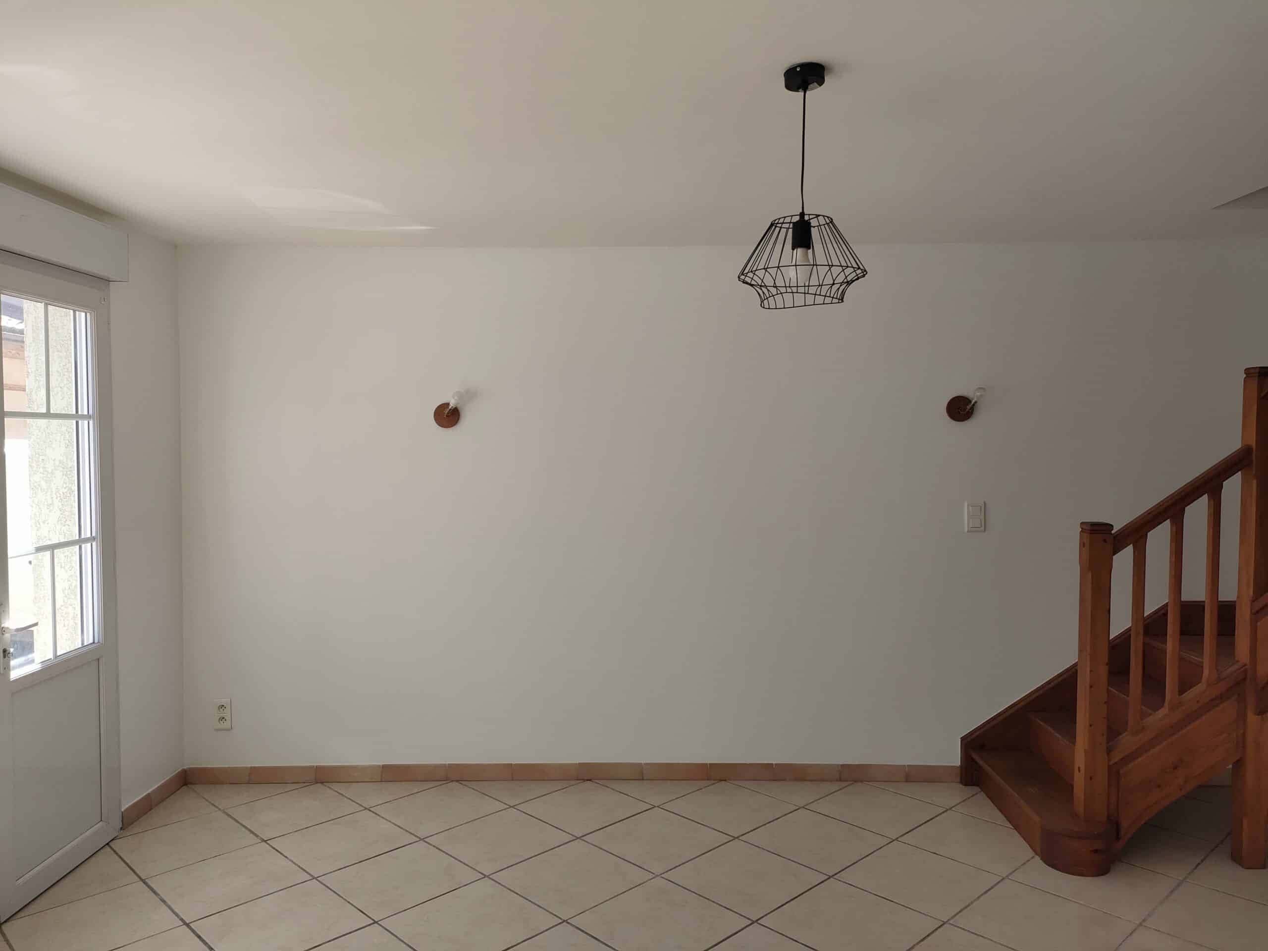 Salon lumineux avec murs blancs, carrelage beige, escalier en bois et luminaire suspendu noir de style industriel.
